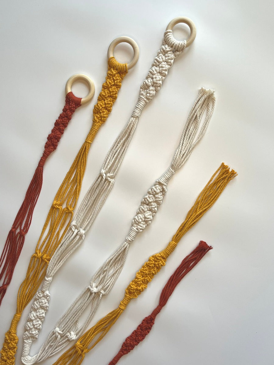 These are the double hat hangers, shown in rust, mustard, and ivory. They have a wooden ring at the top and a braided section at the top, middle, and bottom between the second hat and the tassel. There are square knot details spaced out on the sections that separate to hold the hats. 