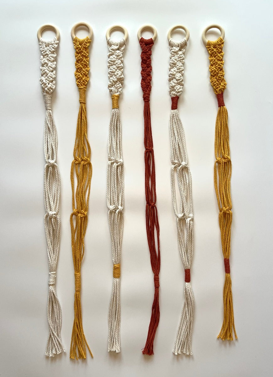 These are the single hat hangers, shown in ivory, mustard, ivory with mustard details, rust, ivory with rust details, and mustard with rust details. They have a wooden ring at the top and a braided section at the top and a tassel at the bottom. There are square knot details spaced out on the sections that separate to hold the hats. 